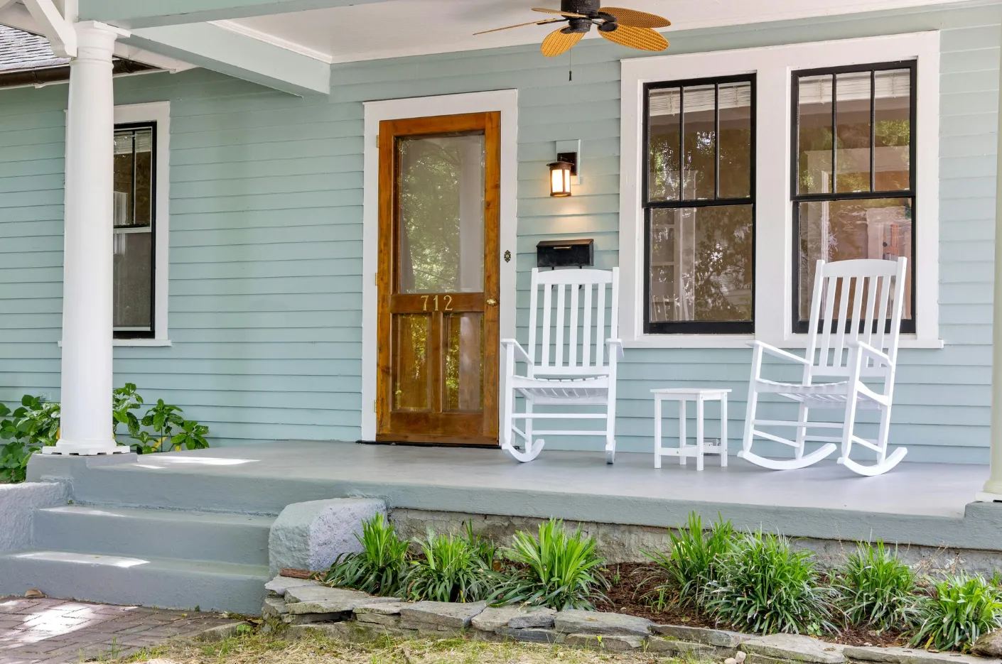 A blue house with a porch. Two white rocking chairs sit near a wooden door.